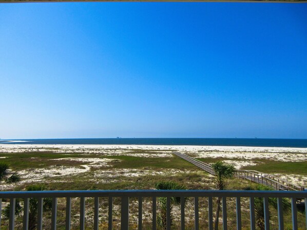A beach view with a wooden boardwalk leading to the water, surrounded by sand and sparse vegetation.