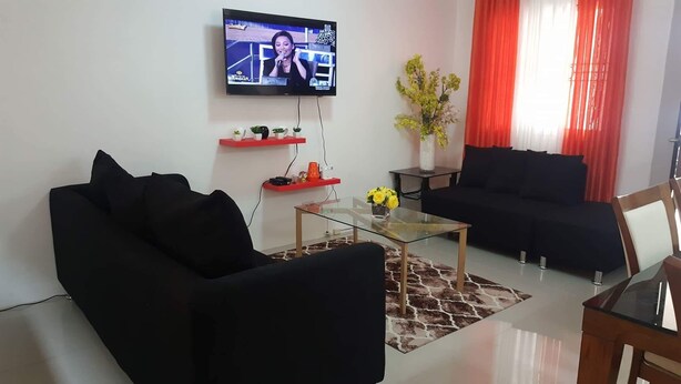 A modern living room with a black sofa, a glass coffee table, and a TV mounted on the wall. - 2 storey townhouse 3 Bedrooms in CEBU FOR RENT - ENTIRE GUEST HOUSE. (Talisay City)