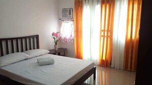 A neatly made bed with pillows, a small bedside table with a vase of flowers, and a window with orange curtains. - 2 storey townhouse 3 Bedrooms in CEBU FOR RENT - ENTIRE GUEST HOUSE. (Talisay City)
