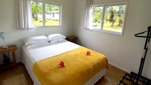 A bedroom with a bed, two windows, a ceiling fan, and a small table with a lamp. - Capture the view, hear the waves -TepaeruVista (Vaimaanga)
