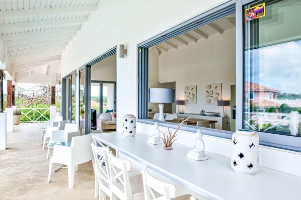 Un espace de restauration extérieur moderne, avec des chaises et des tables blanches, de grandes fenêtres et des vases décoratifs.
