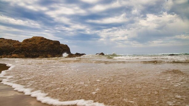 Una playa con olas rompiendo en la orilla y afloramientos rocosos al fondo.