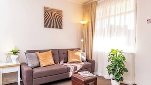 Un salon avec un canapé, deux coussins, une table d’appoint avec une plante, une lampe et une grande fenêtre avec des rideaux.
