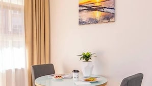 Une chambre d’hôtel avec une table ronde en verre, deux chaises grises, un vase blanc contenant une plante, un tableau représentant un coucher de soleil et une fenêtre avec des rideaux de couleur beige.