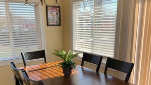 A dining area with a wooden table, six chairs, a potted plant, and two windows with blinds.