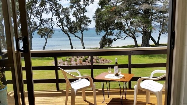 Eine Holzterrasse mit zwei weißen Plastikstühlen und einem kleinen Tisch, mit Blick auf eine Rasenfläche, Bäume und das Meer.