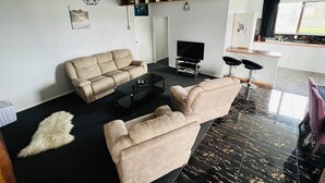 A modern living room with a beige sofa, a black coffee table, and a flat-screen TV. There is a small rug and a white furry object on the floor. - Our house is the perfect holiday home (Raetihi)