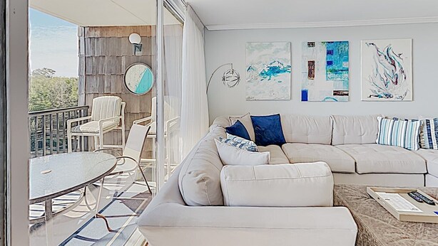 A modern living room with a large sectional sofa, a round coffee table, and a dining area with chairs and a table. There are abstract paintings on the wall and a view of the outdoors through large windows. - Gorgeous, Minutes to Beach, Updated Retreat 414 (Pawleys Island)