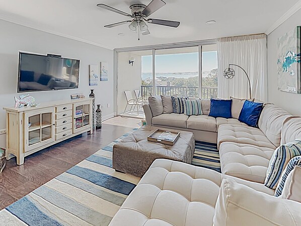 A modern living room with a large flat-screen TV, a beige sofa, a coffee table, and a view of the outdoors through large windows. - Gorgeous, Minutes to Beach, Updated Retreat 414 (Pawleys Island)