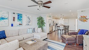 A modern living room with a sofa, ottoman, and coffee table, a dining area with chairs, and a kitchen with appliances. - Gorgeous, Minutes to Beach, Updated Retreat 414 (Pawleys Island)