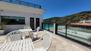 Une terrasse avec des chaises et une table en plastique blanc, donnant sur une plage aux eaux bleu clair et un paysage vallonné.