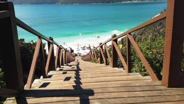 Un escalier en bois mène à une plage bordée d’une eau turquoise, avec des montagnes en arrière-plan.
