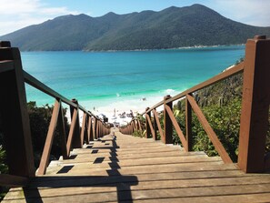 A wooden staircase leading to a beach with turquoise water and mountains in the background. - Loft right on the sand at Prainha in Arraial do Cabo (Arraial do Cabo)