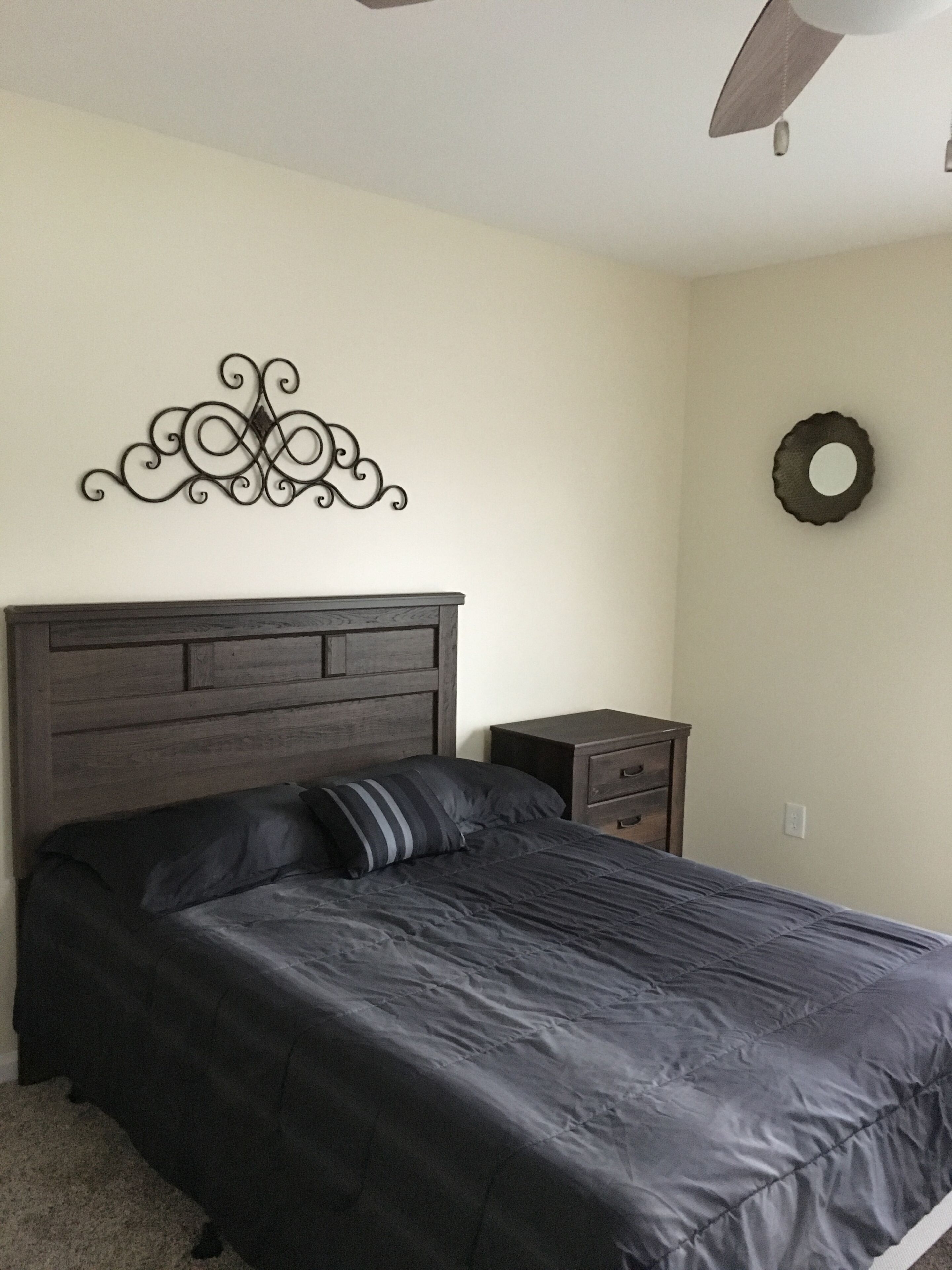 A bedroom with a dark wooden headboard, a bed with a dark gray bedspread, a small bedside table with drawers, and a wall-mounted decorative metal piece.