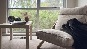 A room with a large window, a cushioned chair, a small table with decorative items, and a carpeted floor.