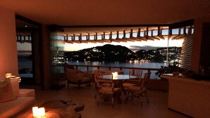 A cozy lounge area with a table and chairs, a round wooden table with candles, and a view of a lake at dusk.