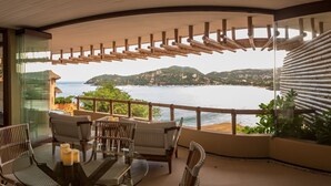 A balcony with a glass door leading to a bedroom, a dining area with chairs and a table, and a view of the sea and hills.