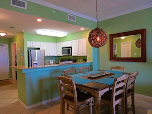 A kitchen with white cabinets, a microwave, and a refrigerator. There is a dining table with chairs and a hanging light fixture. - The Inn at Dauphin Island Unit 103 (Dauphin Island)