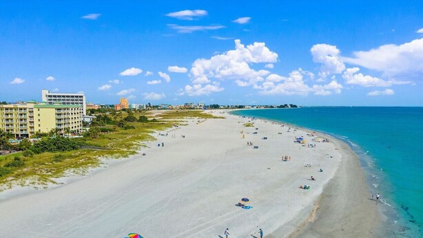 A wide beach with clear blue water, a few umbrellas, and buildings in the background.