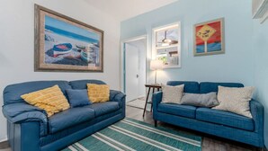 A living room with two blue sofas, a striped rug, a framed painting of boats, and a small table with a lamp.