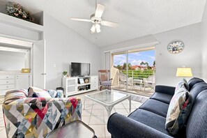 A living room with a television, sofa, and a glass coffee table. There is a ceiling fan, a clock on the wall, and a view of the outdoors through a sliding door.
