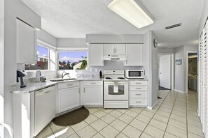 A modern kitchen with white cabinetry, a built-in microwave, and a large window offering a view of palm trees. - Isla Del Sol 2BR Corner Condo w/ Golf Views (Isla Del Sol)