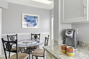 A kitchen with a round glass table, chairs, a coffee maker, and a framed picture on the wall.