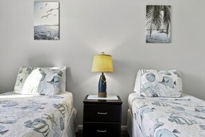 A bedroom with two beds, a nightstand with a lamp, and wall art depicting birds and palm trees. - Isla Del Sol 2BR Corner Condo w/ Golf Views (Isla Del Sol)