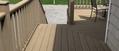 A wooden deck with a railing, a table, and chairs, overlooking a body of water with a grassy area and trees in the background.