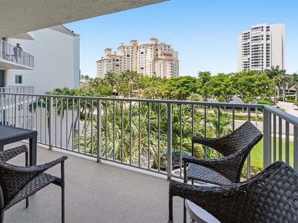 A balcony with wicker furniture, a railing, and a view of tall buildings and greenery. - 1BR Marco Island condo with heated pool, WiFi, and full kitchen. (Marco Island)