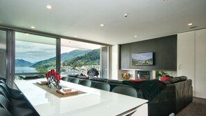 A modern living room with a large dining table, comfortable seating, a television, and a scenic view of a lake and mountains.