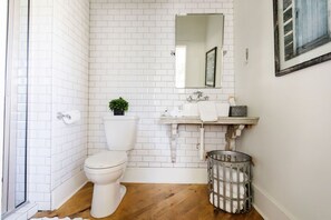 A bathroom with white subway tiles, a white toilet, a wooden floor, a sink with a mirror, and a trash can. - Historical Morrow House Season 5 Fixer Upper, Featured In Cottage Style Magazine (Waco)