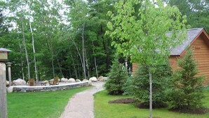 Un chemin de gravier mène à un chalet en bois, entouré d’un mur de pierres et de verdure.