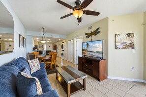 A living room with a sofa, coffee table, TV, ceiling fan, and artwork on the wall. - Sunset Vistas 610N: Top Floor Gulf Front Beauty (Treasure Island)