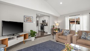 Ein modernes Wohnzimmer mit Flachbildfernseher, einer Couch, einem Couchtisch und einer Küche im Hintergrund.