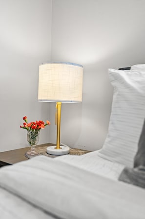Uma cama com roupa de cama branca, uma mesa de cabeceira com abajur e um vaso com flores.