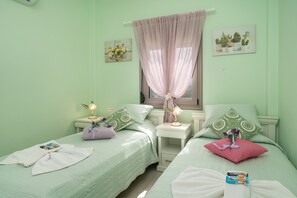 Ein Zimmer mit zwei Betten, jedes mit einem Kopfkissen und einem kleinen Tisch mit Lampe. An den Wänden befinden sich Kunstwerke und ein Fenster mit Vorhang.