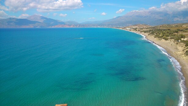An aerial view of a coastal area with clear turquoise waters, a sandy beach, and distant mountains.