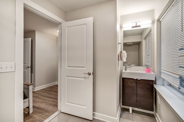 A bathroom with a white door, a sink with a mirror, and a window with blinds.