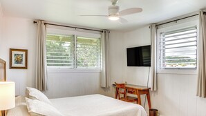 Un dormitorio con cama, escritorio, silla, ventilador de techo, televisor montado en la pared y ventanas con cortinas.