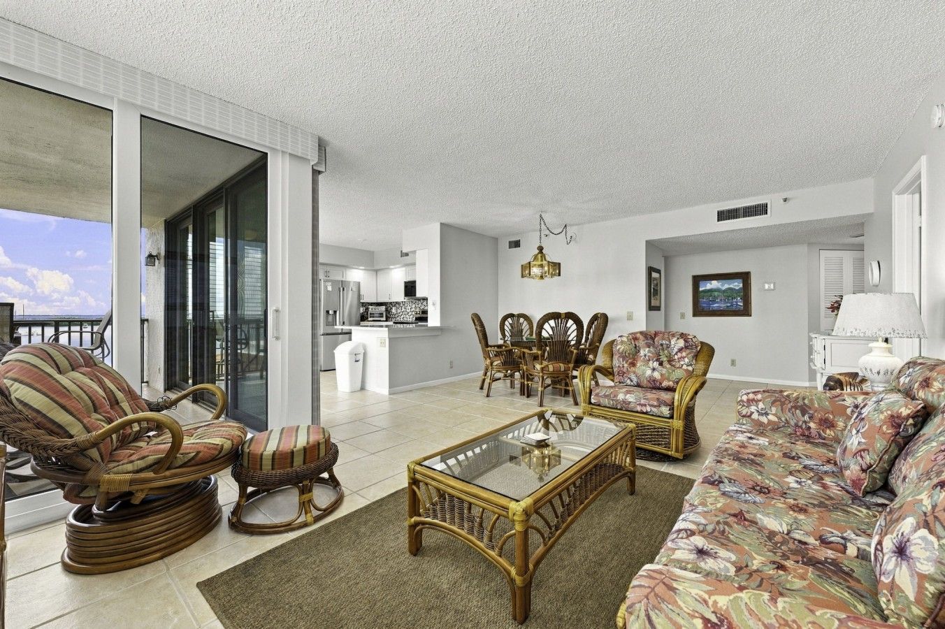 A spacious living area with a balcony, wicker furniture, a glass coffee table, and a view of the kitchen.
