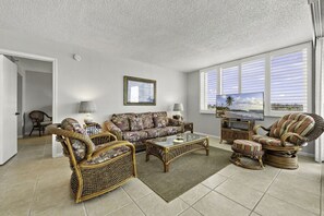 A living room with a sofa, chairs, a coffee table, a television, and a window with blinds. - Bay Front Serenity: 4th Floor 2BD/2BA at Isla Del (Isla Del Sol)