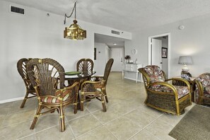 A dining area with wicker chairs and a glass table, a living room with a sofa and a lamp, and a kitchen in the background.