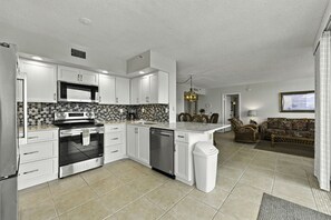 A modern kitchen with white cabinets, a microwave, stove, and dishwasher. There is a dining area with a table and chairs, and a living room with a sofa and a TV. - Bay Front Serenity: 4th Floor 2BD/2BA at Isla Del (Isla Del Sol)