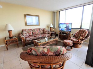 A living room with a floral sofa, wicker chairs, a television, and a large window.