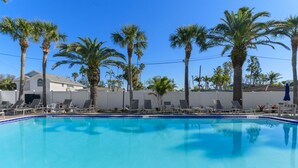 A clear blue pool surrounded by lounge chairs and palm trees, with a white fence and a house in the background.