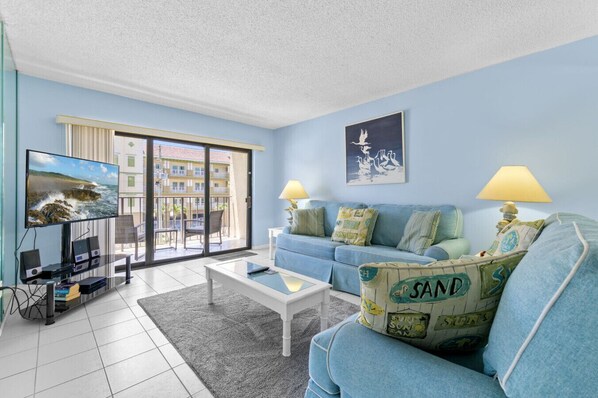 A living room with a blue sofa, a coffee table, a TV, and a balcony with a view of buildings. - Madeira Place 303: 1BD/2BA Retreat Near John`s (Madeira Beach)