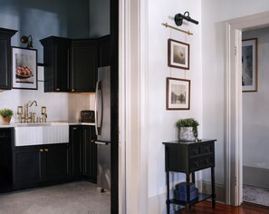 Een keuken met donkere kastjes, een wit aanrechtblad en een koelkast. Er hangen afbeeldingen in lijsten aan de muur en er staat een kleine tafel met een plantje.