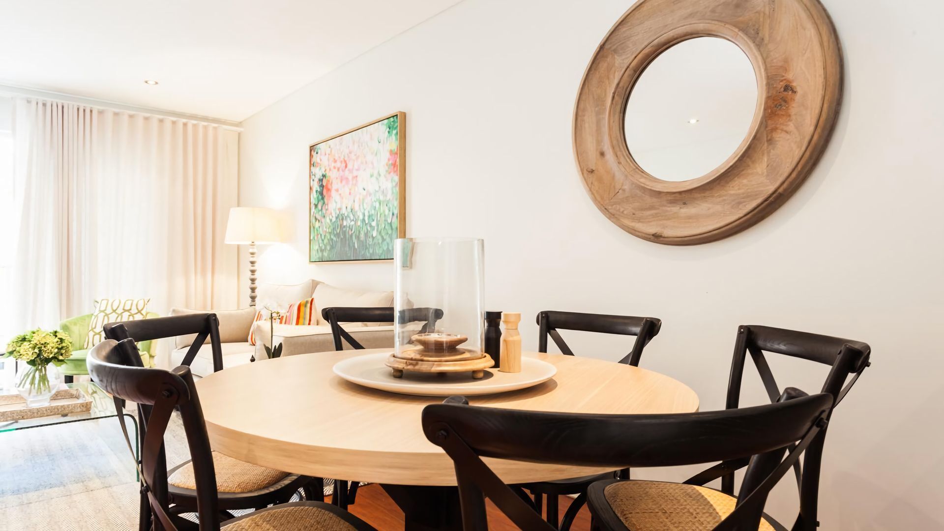 A round dining table with six chairs, a glass-top centerpiece, and a wooden-framed mirror on the wall.