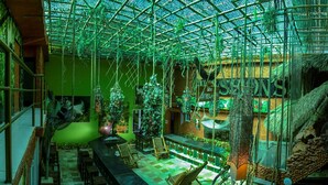 A spacious indoor area with a high ceiling, featuring a long bar, seating arrangements, and a staircase.